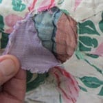 A hand peeling back the outer layer of the Mississippi family quilt, revealing a second older quilt hidden underneath with different patterned cotton fabrics.