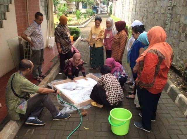 Indonesian textile professionals gathered outdoors around a shallow washing tray during a wet-cleaning demonstration in Jakarta.