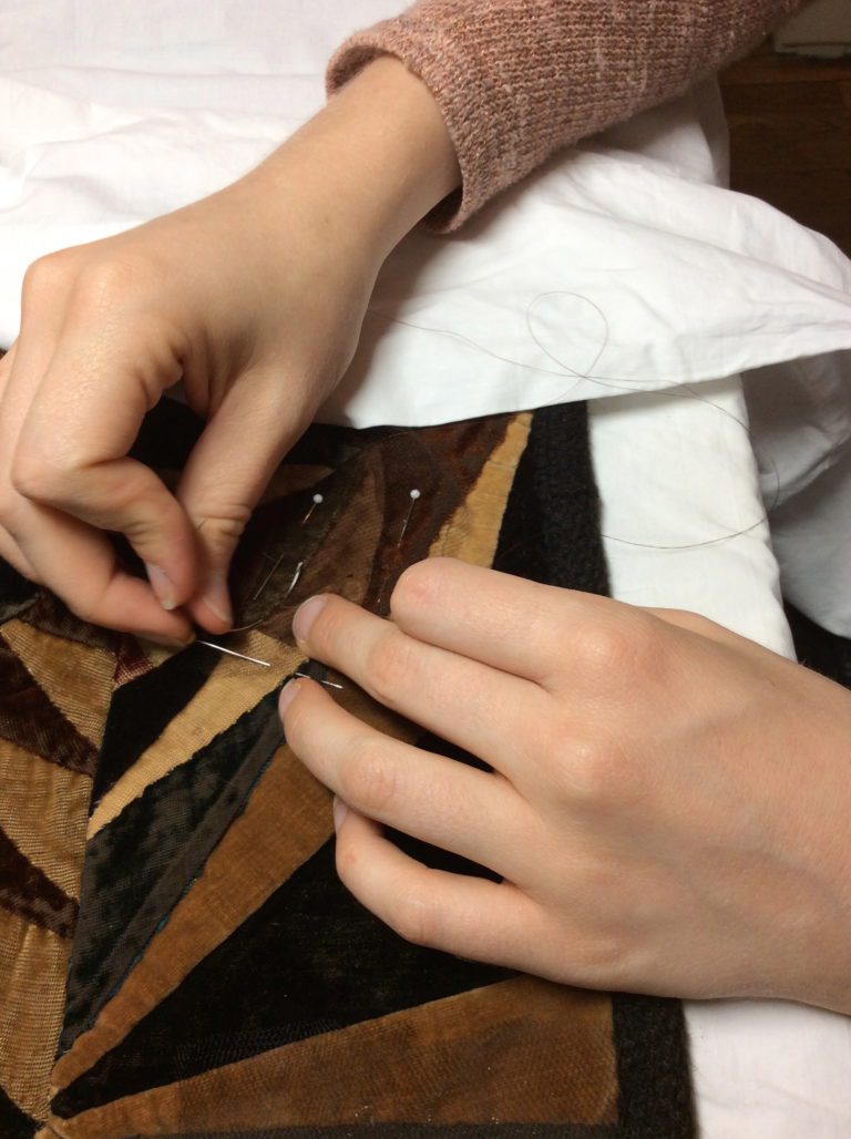 Hands stitching a fragile patchwork textile with a fine needle and thread, supported on white tissue paper.