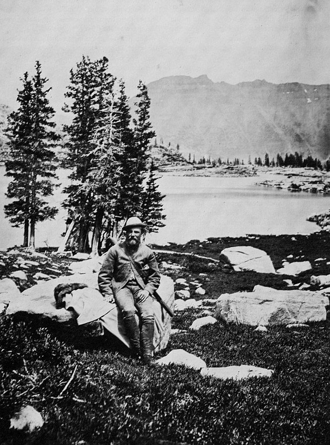 19th-century black-and-white photograph depicting a man seated on a rock in a mountainous landscape.