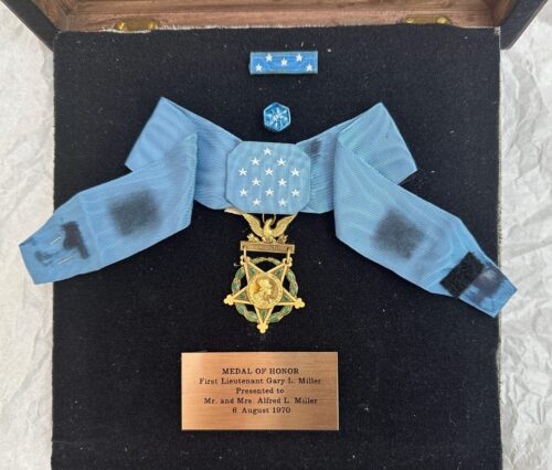Front view of a Medal of Honor suspended from a light blue grosgrain ribbon with white stars, gold medal pendant, and engraved plaque identifying First Lieutenant Gary L. Miller, dated August 6, 1970.