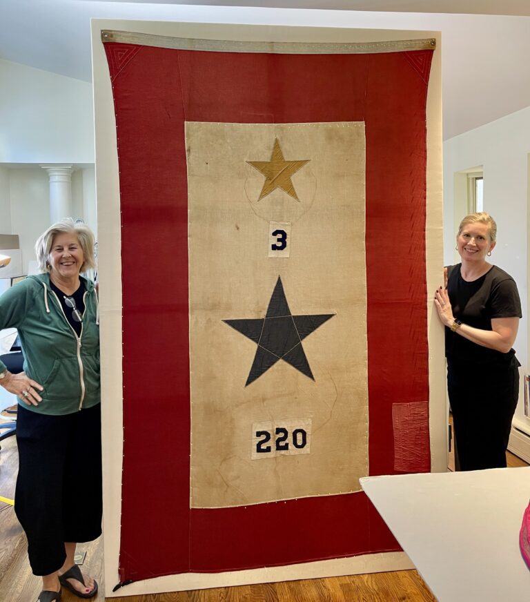 Julia and colleague stand on either side of a large historic service banner featuring a gold star above a black star and the numbers “3” and “220,” shown after conservation treatment and mounting.