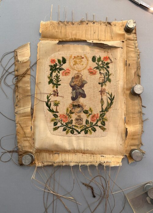 Antique silk embroidery of Mary featuring floral motifs and a central figure, secured to a larger fabric support with pins and weights during treatment.
