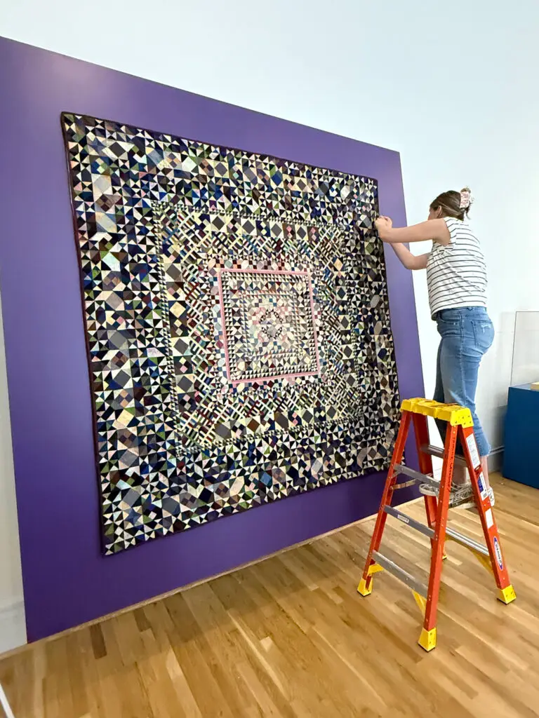 Repair of antique rug hanging on purple wall with textile worker standing on orange ladder.