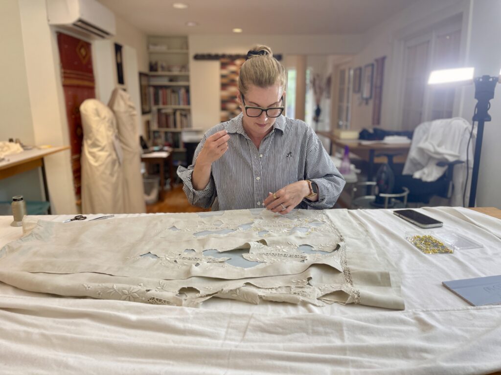 White woman wearing blue blouse and black glasses, sits at table repairing s cream colored linen cloth in front of her. One hand up, one hand down, as she sews.