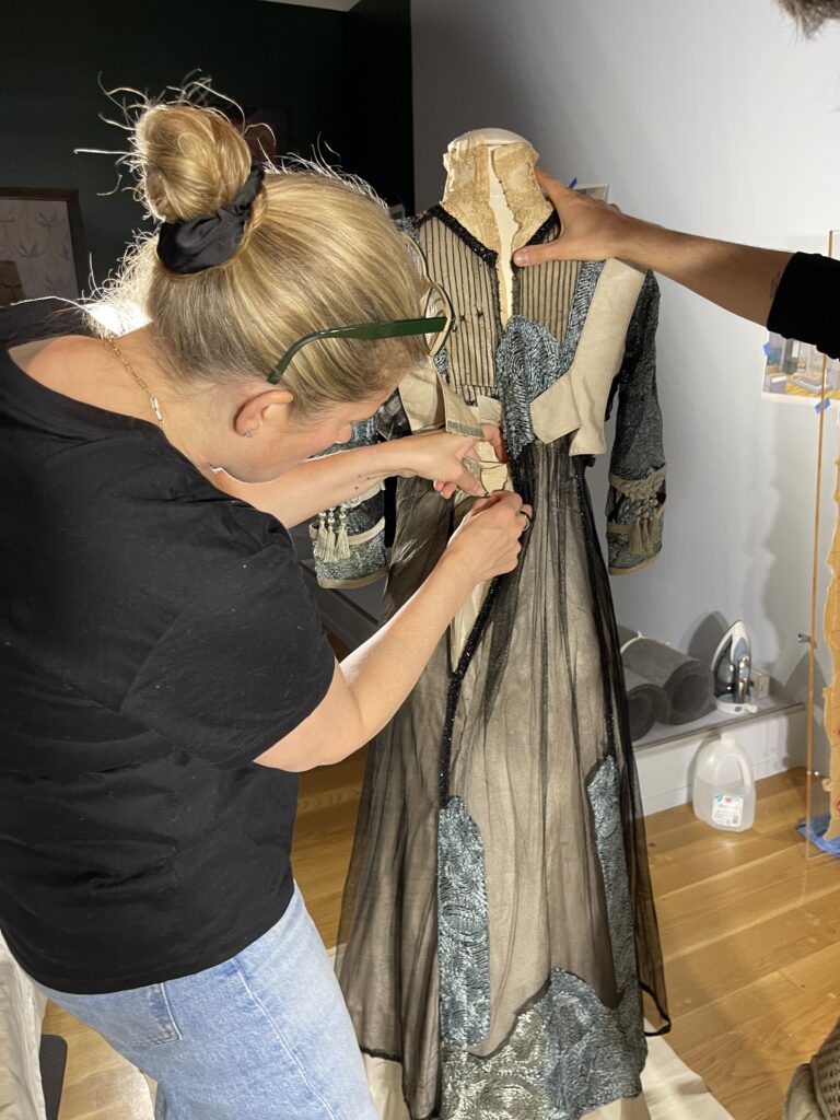 Same white woman in black shirt attaches hooks and eyes in the back of a blue and black long dress.
