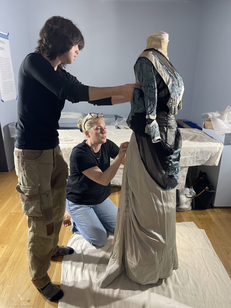 Young man in black shirt standing on left and white woman in black shirt kneeling are holding a dress form with a blue and black dress, and attaching the closures on the back of dress.