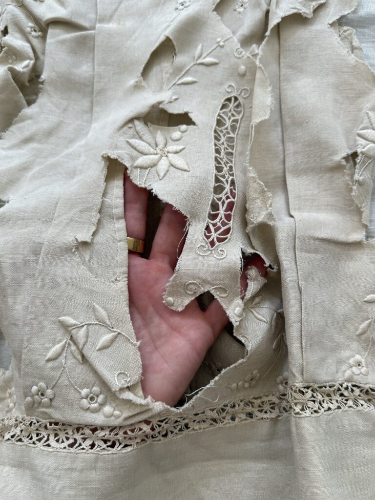 Cream colored linen with cream colored floral embroidery and lace inserts, with large holes cut into fabric in center, where a hand is visible.