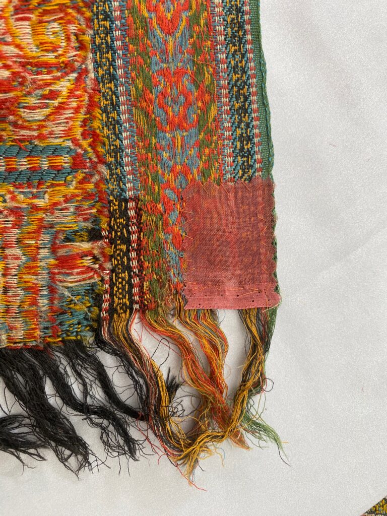 Detail of red, blue, green fringed woven striped cloth with pale red patch in bottom right corner.