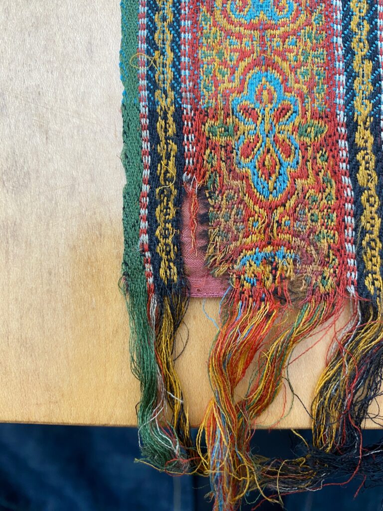 Detail of same red, blue, green fringed woven striped cloth from the other side, showing just a tiny bit of the pale red patch repair.