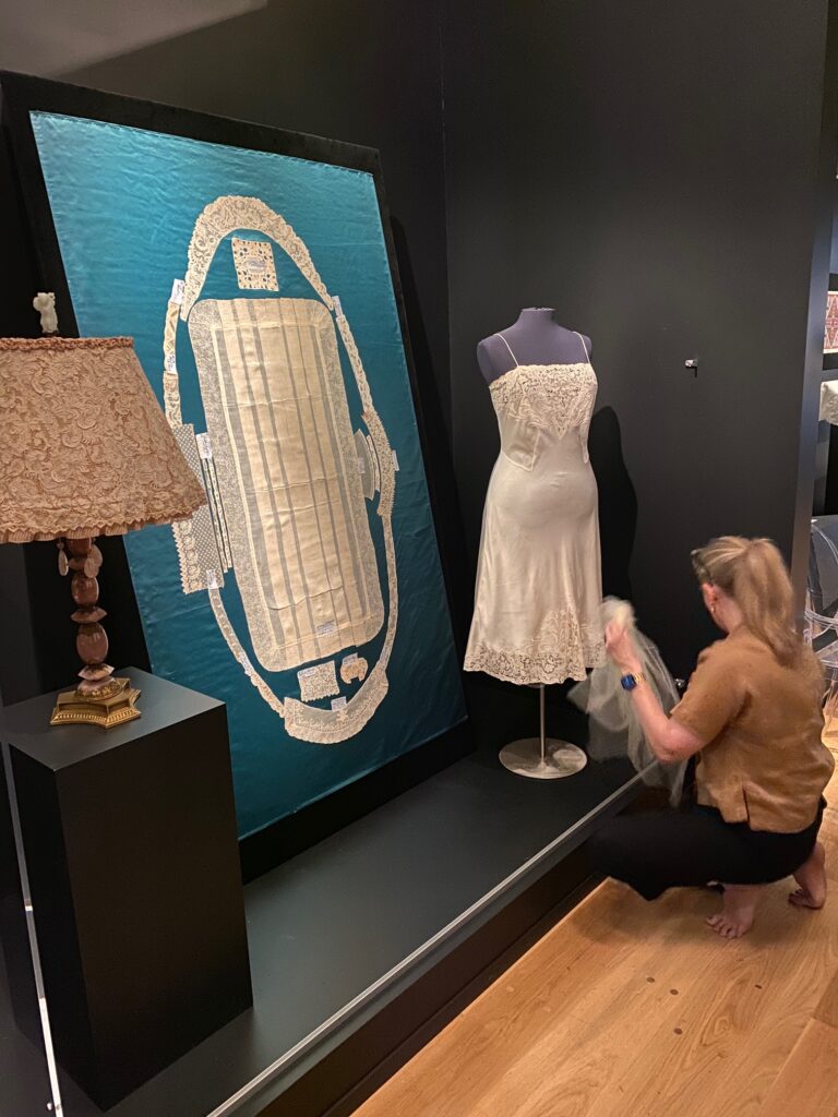 Woman with blonde hair is squatting in front of a white silk lace slip adjusting the hem. A large vertical turquoise colored silk panel with white lace pieces on it is to the left.