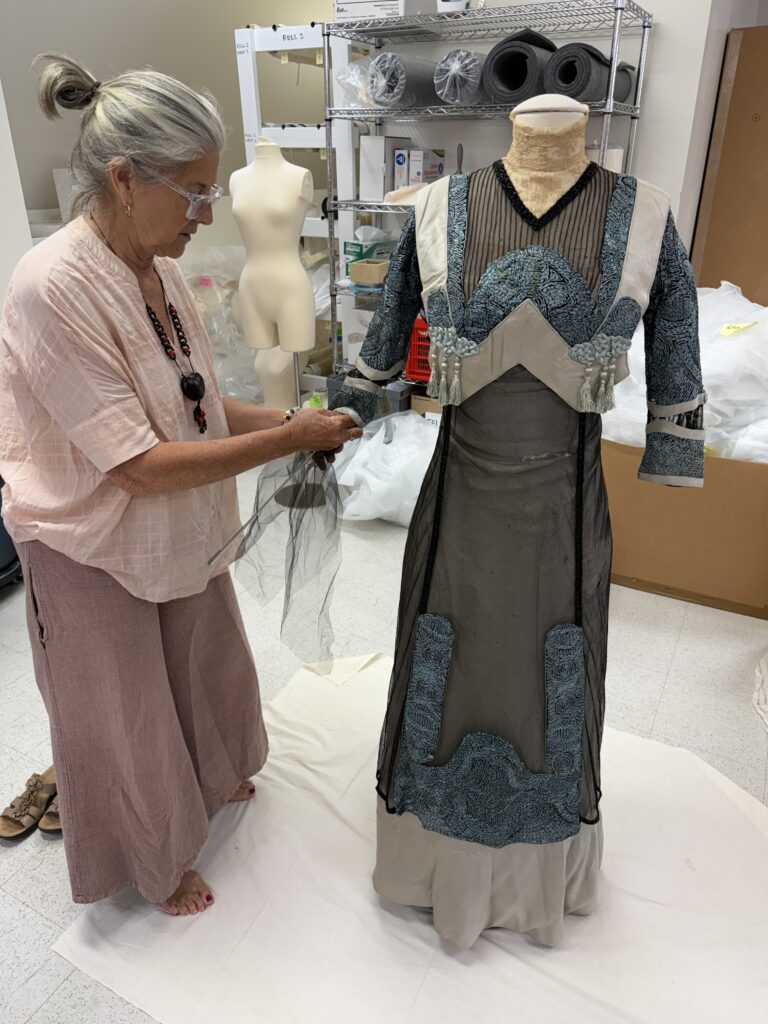 Woman with gray hair, pink blouse, brown trousers holding black netting in both hands, as she inserts it into the left sleeve of a blue and black lace and silk embroidered long dress.