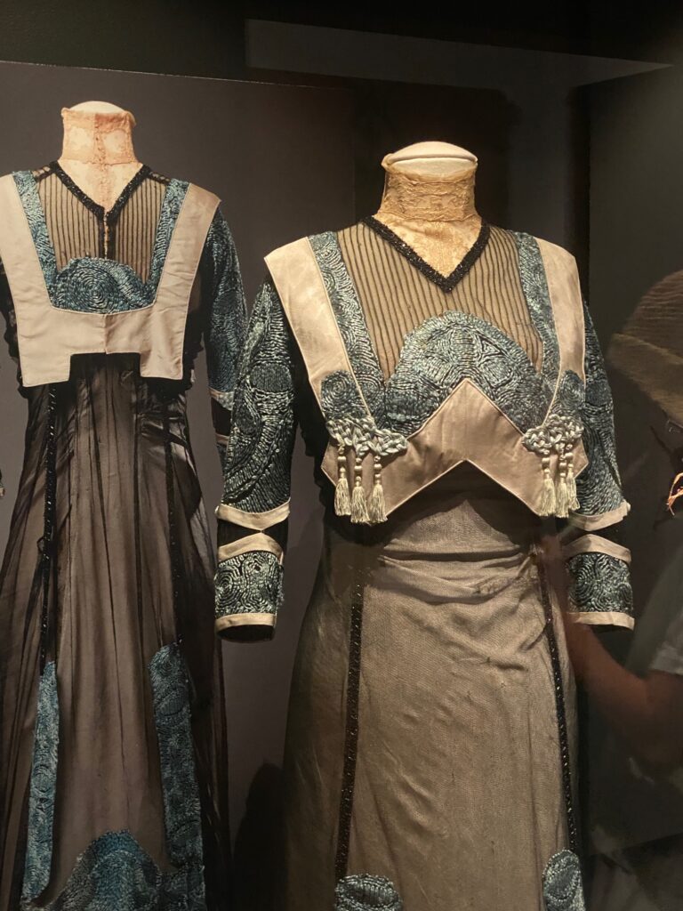 Long formal dress on a dress form; the back view is on the left, the front view is on the right. Blue silk embroidery, black netting lace, V-shaped bodice with tassels, high lace collar, long blue silk embroidered sleeves.