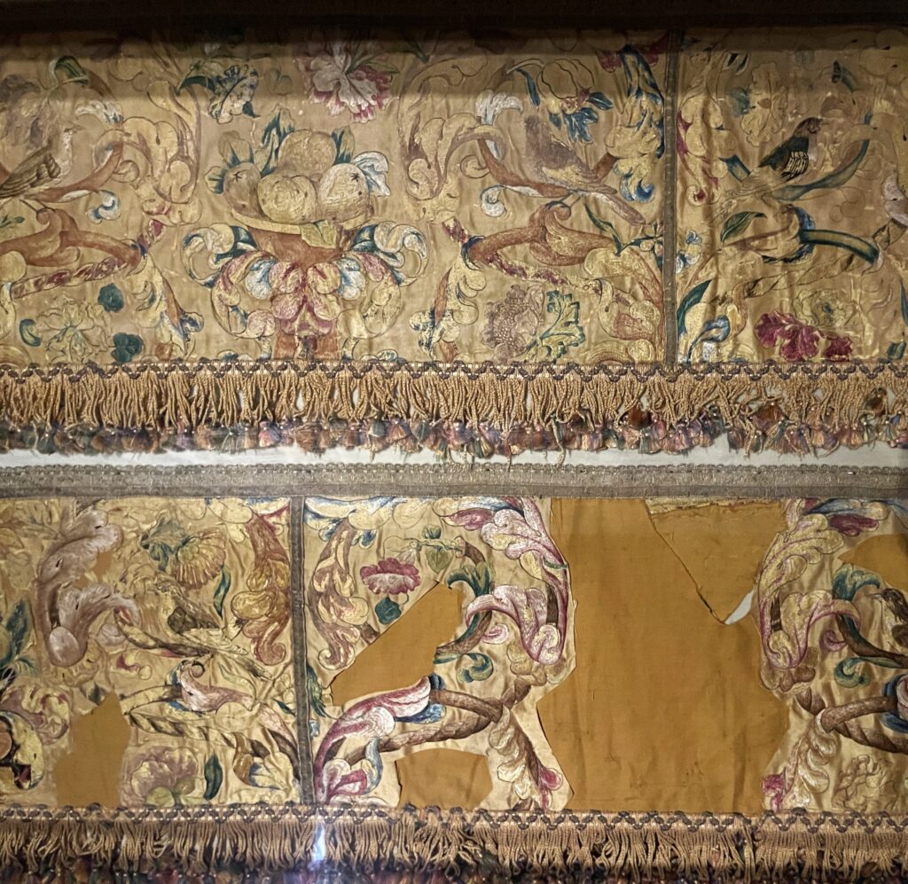 Two textiles shown horizontally. The top one is yellow gold shiny embroidery with tassels along the bottom. The textile below it is similar in color and embroidery, but there are three large areas of solid yellow fabric, where the embroidery is missing.