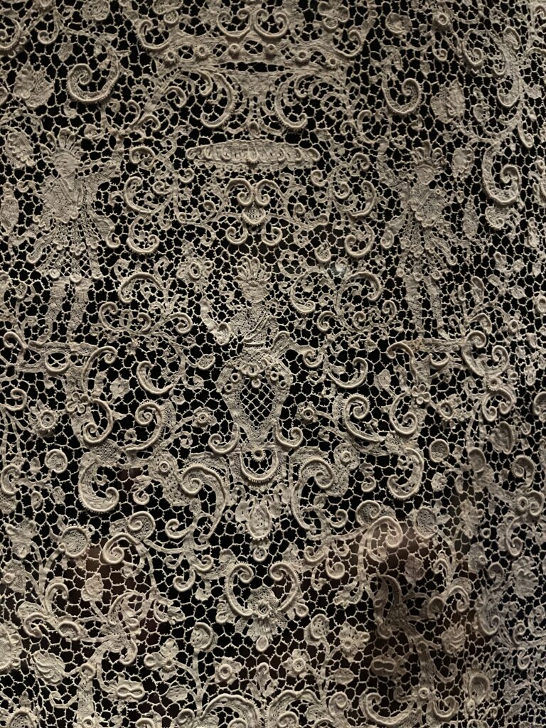 Close detail of very fine intricate lace depicting a central figure and floral patterns on a dark background..