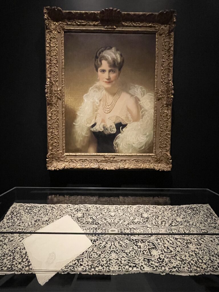 Portrait of Marjorie Merriweather Post; Gold framed painting of white woman wearing pearls and fluffy white lace, with large panel of intricate white lace on display below portrait.