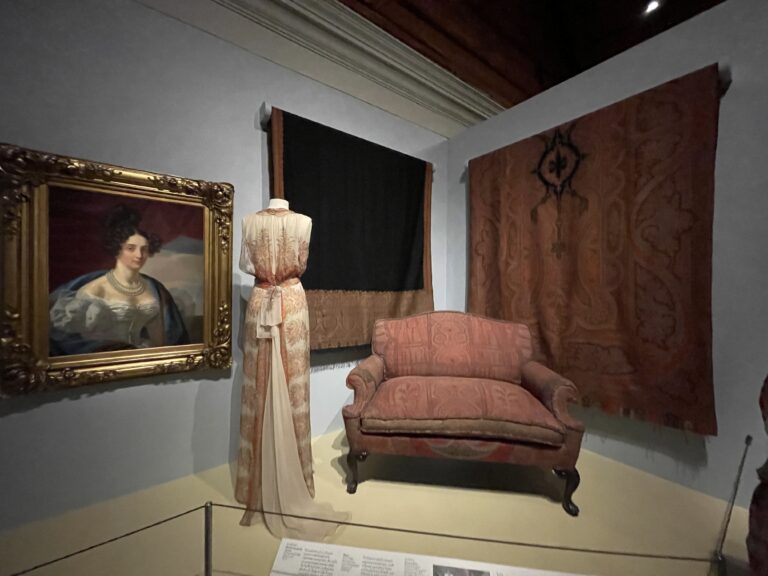 A corner of a room with a painting of a woman in white silk bodice; a tall dress form with a pink and cream-colored long dress; a large black cloth on left wall, a large red colored cloth on right wall, a small red colored sofa in front.