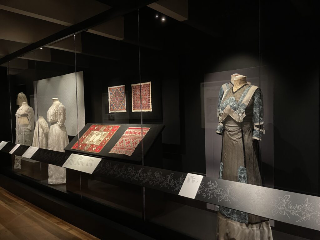 Dark room with black walls displaying four standing dress forms with historic clothing, and four flat red colored fabrics.