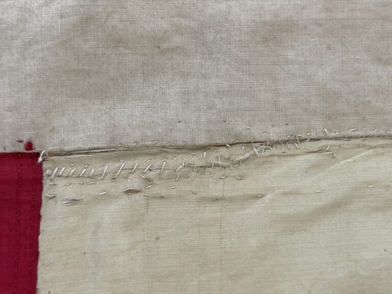 Close-up of a hand-stitched seam on beige fabric with a red edge at the bottom.