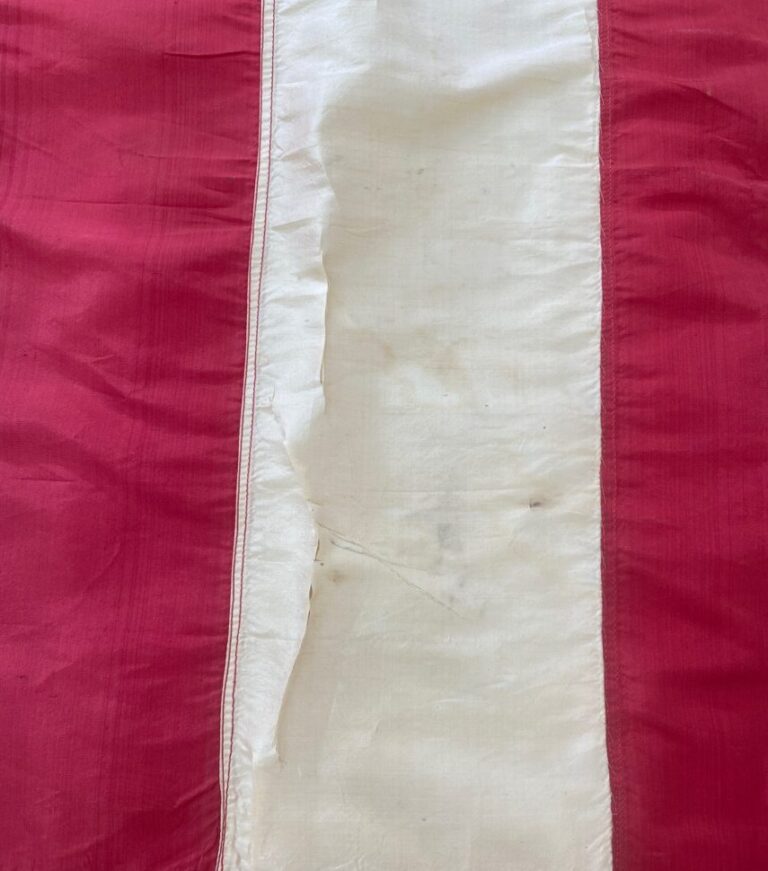 Detail of long narrow vertical white shiny silk strip on red background, with holes and tears.