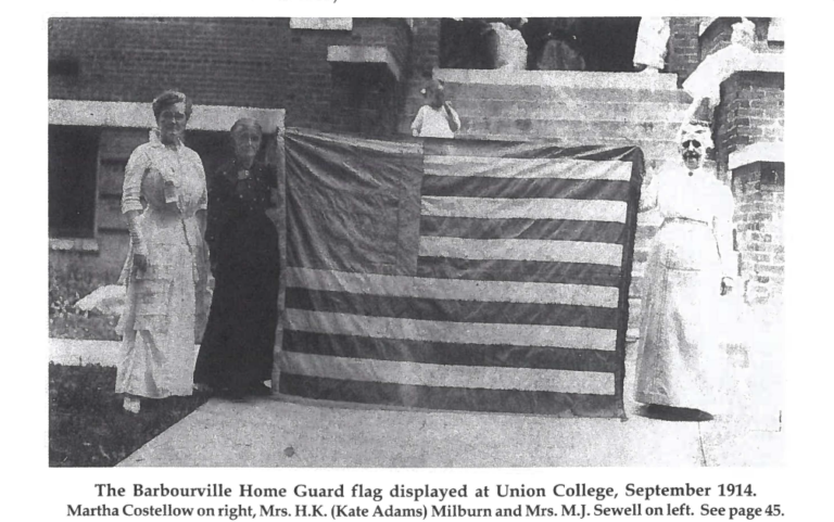 Black and white photo of three women, dressed in 1914 attire, standing in front of a brick building. They are holding a flag designed with horizontal stripes and a solid-colored dark block in the top left corner, which awaits its stars.