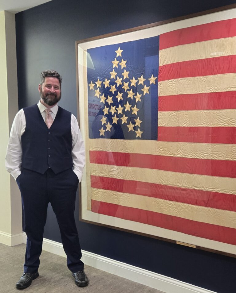 Bearded man in a navy vest and suit stands with hands in his pockets beside a large framed American flag artwork.