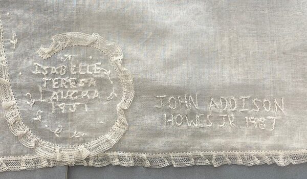 Detail of white cotton dress with lace trim includes two names and dates embroidered in white thread, honoring those christened in this dress.