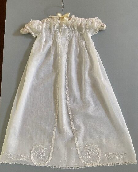 White long-sleeved christening dress with puff sleeves, lace bodice and ornate trim around the hem. Family names are embroidered in white thread along the hemline.