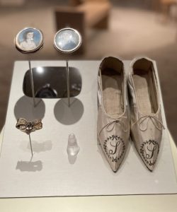 Mother Seton's ivory silk slipper displayed on a white plinth alongside two miniature portrait medallions, a decorative brooch, and a small translucent bottle in a museum installation.