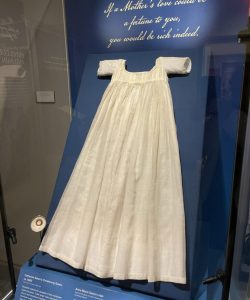 Cream colored christening gown mounted on a blue display panel inside a glass museum case.
