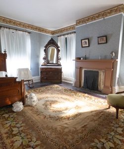 Frederick Douglass's bedroom with large Victorian bed, and decorative bed pots sits at foot of bed. Aubusson-style floral carpet lays on hardwood floors. The room has a fire place, light blue walls, a light green, tufted Victorian-era chair and white curtains.