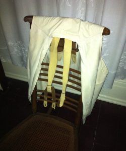 Frederick Douglass light beige waistcoat and cream-colored suspenders draped over ladderback chair in room with white embroidered curtain in background.