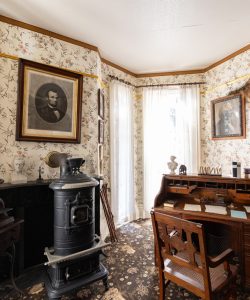 Frederick Douglass's office with portraits of Presidents on flowered wallpaper, a wooden desk with drawers and cubby holes. A black iron woodburning stove sits to the left of the desk. White curtained windows are in front of the desk.