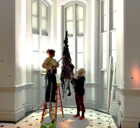 Two textile conservators carefully clean a suspended fiber sculpture in a gallery space.