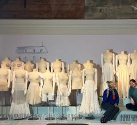 Julia and her colleague seated beside a display of dress forms wearing historical undergarments in a museum gallery.