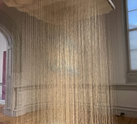 A large hanging installation composed of hundreds of fine vertical fiber strands descends from a rectangular frame, forming a translucent curtain within the gallery.