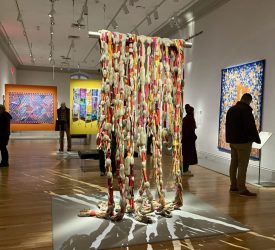 Large-scale hanging fiber sculpture installed in the center of the gallery, surrounded by quilts and visitors viewing the exhibition.