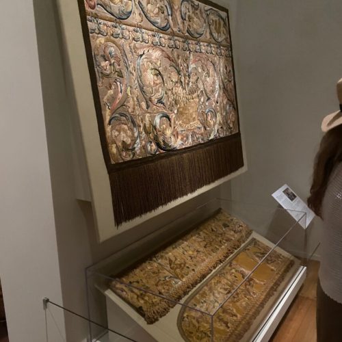 View of textiles on display. Top of the image, a gold-colored shiny textile with green and red curvilinear embroidery patterns, with dark brown edges and long tassels hanging from the bottom. Below in the bottom of photo are two long textiles seen from a side angle, laying on a tan colored angle. The two pieces of fabric are long, narrow, yellow in color.