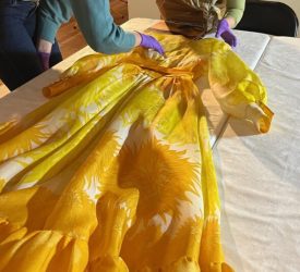 Two textile conservators carefully examine a vibrant yellow gown with bold floral motifs from Princess Grace Kelly's collection during exhibition preparation.