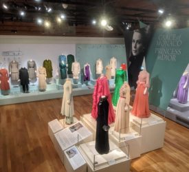 Exhibition gallery at Marjorie Merriweather Post’s Hillwood Estate displaying dozens of Princess Grace Kelly’s dresses and ensembles on mannequins. The show, titled Grace of Monaco: Princess in Dior, features formal gowns and tailored outfits arranged on raised platforms beneath gallery lighting.