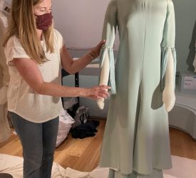 A textile conservator adjusts the sleeve of a mint-green dress displayed on a mannequin in a gallery setting.