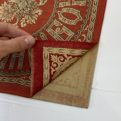 Red and cream-colored printed cloth handkerchief, with bottom right corner lifted up to show brown board below.