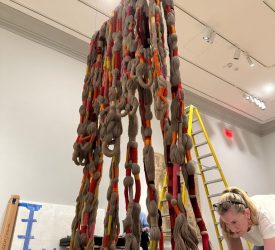 Conservator kneels on the gallery floor adjusting the pooled strands of a large suspended rope installation.