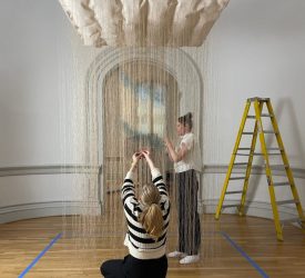 Two textile conservators carefully align hanging strands of a suspended fiber installation in a museum gallery.