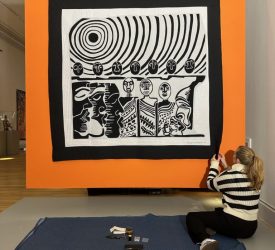 Conservator adjusting large quilt on orange gallery wall.