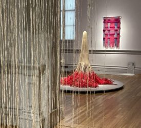 Fiber sculpture with cascading cords and red rope displayed in gallery.