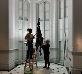 Two textile conservators carefully clean a suspended fiber sculpture in a gallery space.