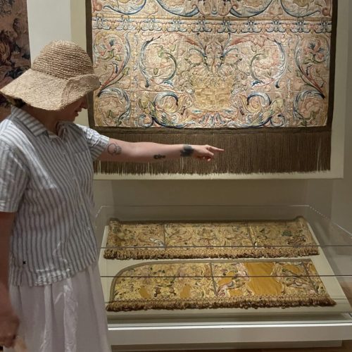 Woman in straw hat, blue striped blouse and white skirt points to three golden colored embroidered textiles on display, to her right.