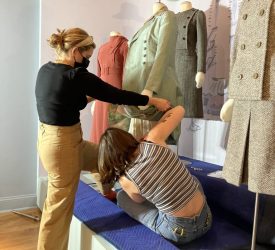 Two conservators carefully adjust the sleeve and positioning of a mint green coat ensemble on a mannequin during installation of Princess Grace Kelly’s garments.