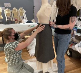 Two conservators carefully adjust the waistband and fit of a gray skirt on a padded dress form during installation of Princess Grace Kelly’s garments, surrounded by protective tissue and mount materials.